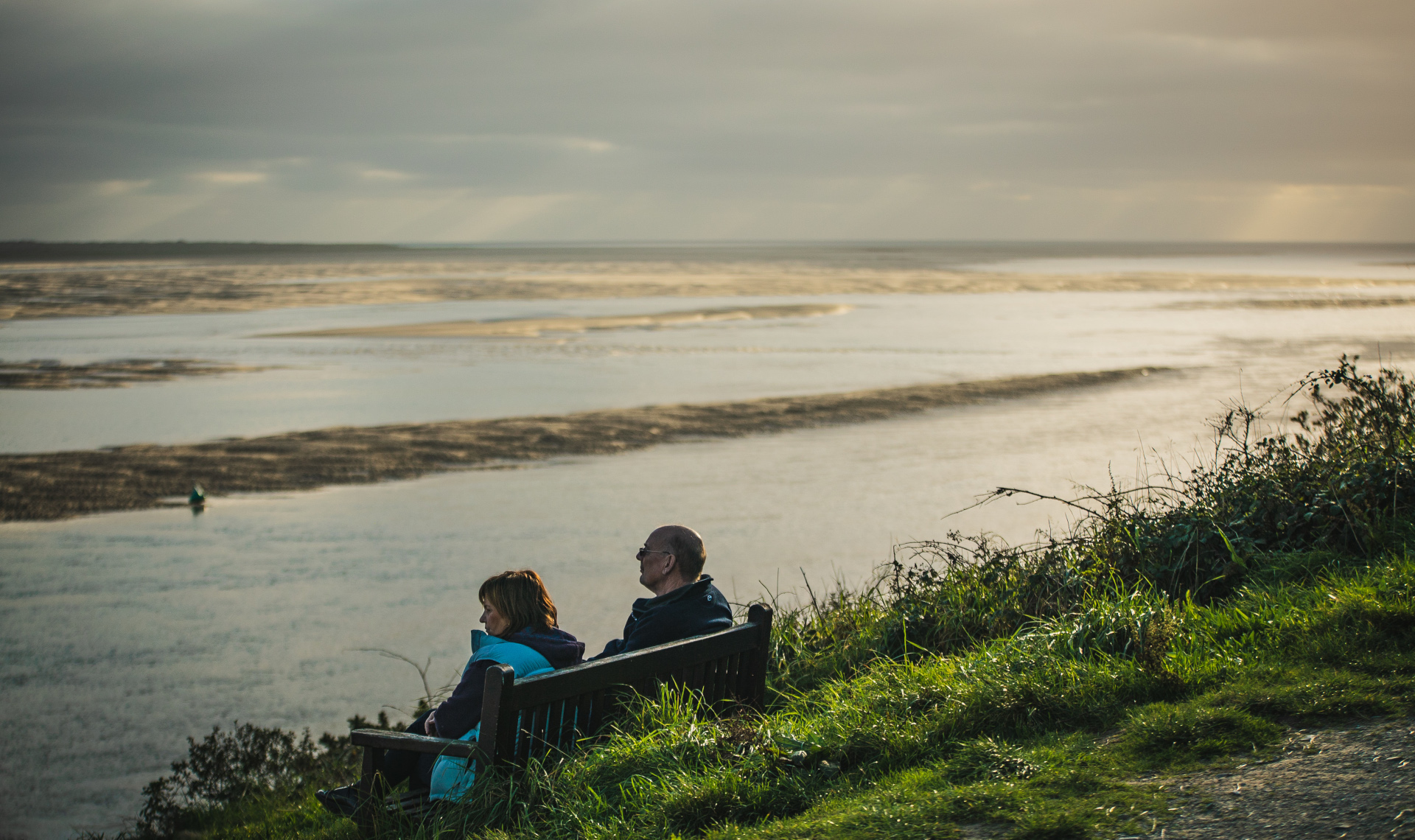 An elderly couple sitting on a wooden bench looking out to sea
